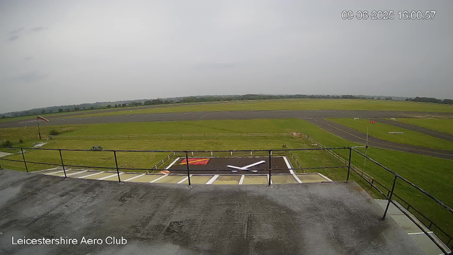 A view from a webcam at Leicestershire Aero Club showing a grassy airfield with runways stretching into the distance under a cloudy sky. In the foreground, there is a helipad marked with a large white "H" and a red square, surrounded by a black railing. The date and time are displayed in the top right corner as "09-06-2025 16:00:57."