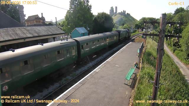 A green passenger train with multiple carriages is stopped at a small railway station platform. The platform has a green bench and flower pots, as well as a wooden fence with wire mesh. To the right, there is a tall wooden telegraph pole with electrical wires, and a narrow path runs alongside the station. In the background, a hillside with a castle ruins or historic structure is visible, surrounded by trees and greenery. The weather appears sunny with clear skies. The date and time displayed are June 9, 2025, at 17:00:53.