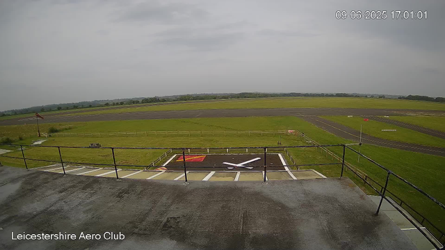 A view from a webcam at Leicestershire Aero Club showing an empty helipad with a large white "H" and a smaller red square with a yellow border. The helipad is surrounded by a metal railing, and beyond it, there are grassy areas and a runway stretching into the distance under a cloudy sky. The timestamp in the upper right corner reads "09-06-2025 17:01:01."