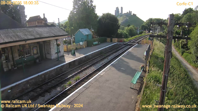 A small train station platform with a stone building and a bench on the left side. Two train tracks run through the center, with some grass growing between the rails. On the right side, there are overhead electrical wires and poles, with a narrow pathway beside a lush green hedge. In the background, a hill with castle ruins or old stone structures is visible, surrounded by trees, under a clear sky with the sun shining brightly. The date and time are displayed in the top left corner.