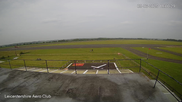 A view from a webcam at Leicestershire Aero Club shows an outdoor area with a helipad featuring a large white helicopter symbol painted on the black surface. The helipad is enclosed by a white fence, and there is a small red and white wind sock on a pole nearby. Beyond the helipad, there are grassy fields and a runway, with two people walking in the distance under a cloudy sky. The image is timestamped "09-06-2025 18:00:29."