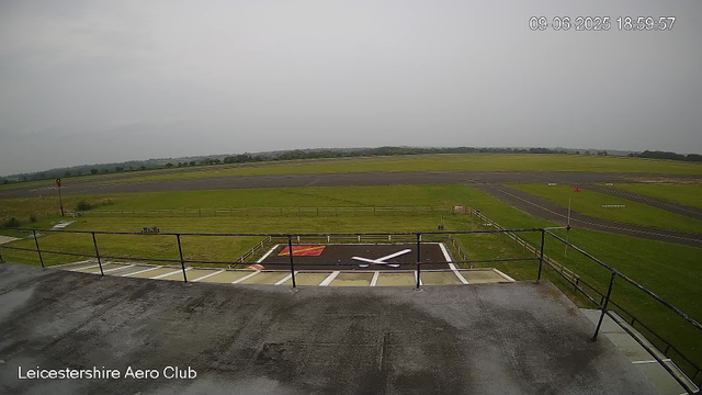 An empty helicopter landing pad with a large white "X" on a dark surface, surrounded by a small white and red border. The pad is located on a raised platform with black railings. Beyond the platform, there are grassy fields and a runway in the background under a cloudy, overcast sky. The location is Leicesterhire Aero Club, as indicated by the text at the bottom left corner of the image. A timestamp shows the date and time as 09-06-2025 18:59:57.