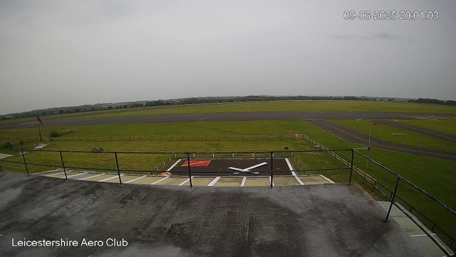 An outdoor view from a webcam at Leicesterhire Aero Club shows a cloudy sky over an open grassy airfield with a runway in the distance. In the foreground, there is a helicopter landing pad with a large white "X" marking its center, surrounded by a fenced area. The scene appears to be on a rainy day, as the surface in the foreground is wet. The timestamp reads "09-06-2025 20:01:03."