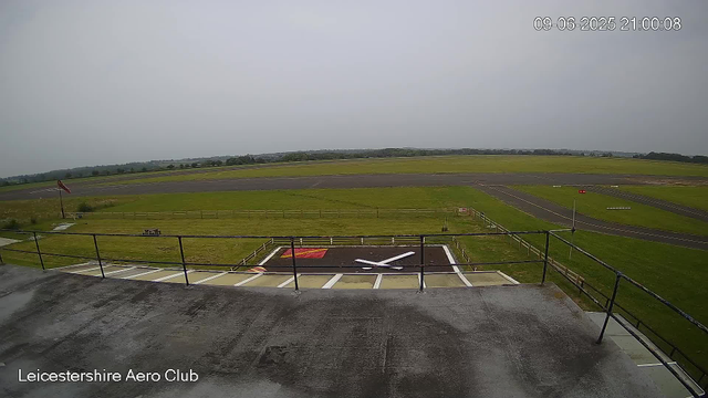 An empty helipad with a large white "X" painted on it, surrounded by a black border. There is a red and yellow marking next to the "X." The helipad is located on an elevated platform with a metal railing in the foreground. In the background, there is a grassy area with a flagpole, and beyond that, an empty runway stretching into the distance under an overcast sky. The image is from the Leicestershire Aero Club, and a timestamp in the top right corner shows the date and time as September 6, 2025, at 9:00 PM.