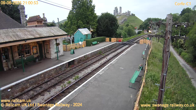 An empty train station platform with a small blue building, several green benches, and a row of potted plants. Railway tracks run alongside the platform, with a lush green hillside and the ruins of a castle visible in the background. There are utility poles with wires along the right side of the platform, and a small green hedge and a yellow fence at the far end of the station. The sky is overcast. The image has timestamps and website information bar overlays.
