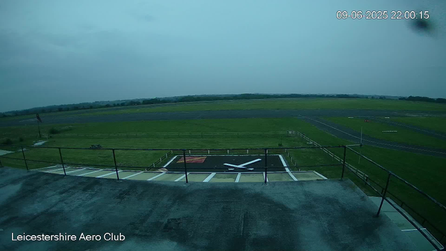 The image shows an overcast sky at the Leicester Aero Club with a grass airfield and a helipad marked with a white "X" in the foreground. A flag is fluttering on a pole on the left side of the image, and there are some small structures and pathways on the grass. The timestamp on the upper right corner indicates the date and time as September 6, 2025, at 22:00:15.