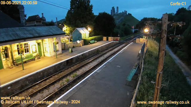 A small railway station platform at dusk, illuminated by warm yellow lighting. There are green benches and a payphone box on the platform. Tracks run alongside the platform, with a grassy area and a dirt path on the right. A tall wooden utility pole with electrical wires extends along the platform’s edge. In the background, a green building with a corrugated roof and hanging flower baskets is visible, along with a scenic hillside featuring castle ruins atop a hill, surrounded by trees and greenery. The sky is cloudy and darkening as night approaches.