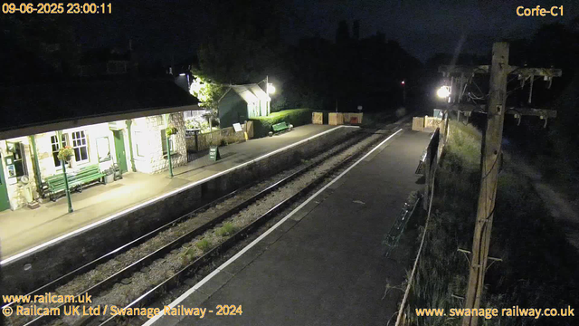 A nighttime view of a small railway station platform with a stone building on the left, featuring green benches and hanging flower baskets. The platform is lit by a few street lamps, and there are some signs and a small shed further down the platform. The railway tracks run through the middle of the image, with barriers and barriers on the far end. To the right, there are wooden utility poles with wires and some fencing. The scene is dark, and the overall atmosphere is quiet and empty.
