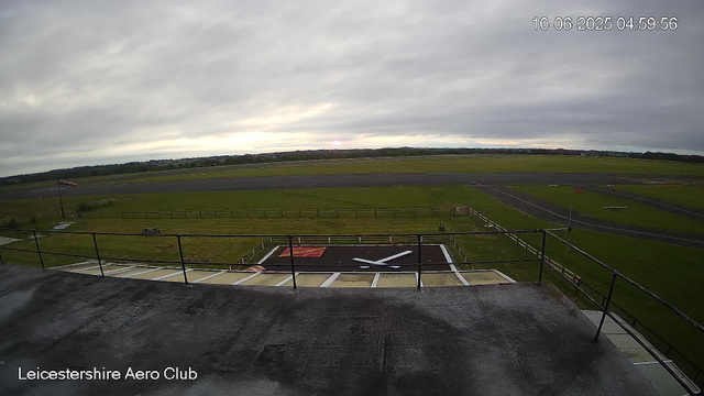 A view from a webcam at Leicestershire Aero Club showing an empty aircraft parking area with a large black helipad marked with a white helicopter symbol in the foreground. Beyond the helipad, there is a grassy field, a small windsock, and a runway in the distance with a cloudy sky overhead. The timestamp in the top right corner reads October 6, 2025, at 04:59:56.