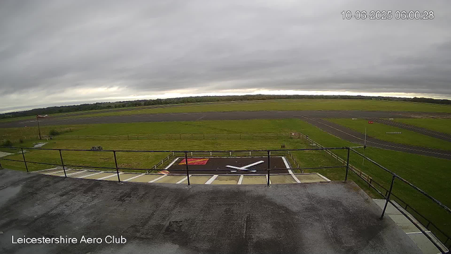 Overcast sky view of an airfield from a webcam, showing a tarmac runway and a grassy area with a windsock on a pole in the foreground. The image includes a date and time stamp (10-06-2025 06:00:28) in the top right corner and the label "Leicestershire Aero Club" in the bottom left corner.