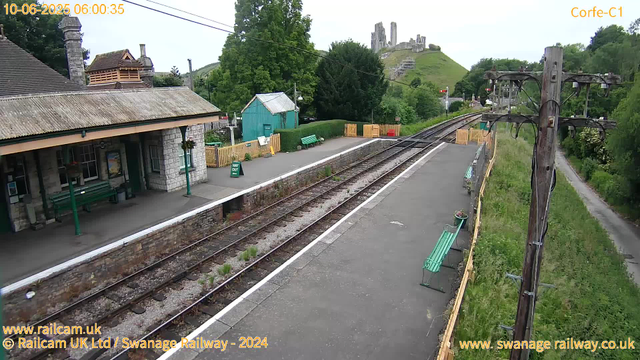 A small train station platform with a stone building featuring a tiled roof. There are green benches, some potted plants, and a yellow fence around the platform. To the right, there are overhead power lines and a pole. The railway tracks run through the center, with greenery and trees on either side. In the background, a green hill with ruins of a castle or fortress is visible. The time and date are displayed in the top left corner, and there are website URLs at the bottom of the image.