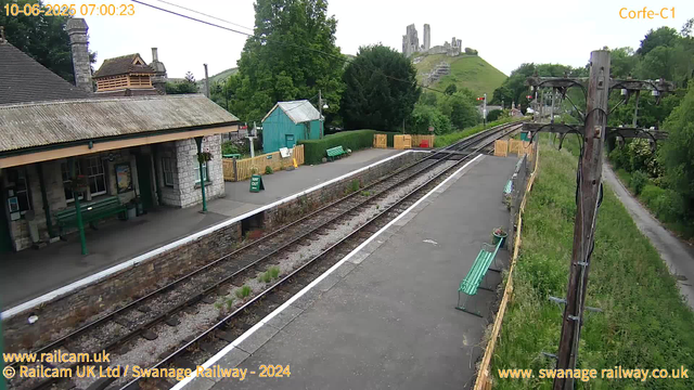 A small train station platform with green benches, a stone building with a sloped roof on the left, and a green hut in the background. Railroad tracks run through the center, with trees and a hill topped by ruins in the distance. A wooden utility pole with wires is on the right, and there is a pathway with a fence along the platform. The sky is overcast, indicating early morning.