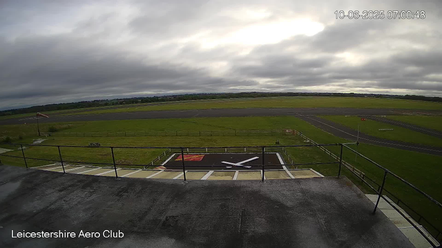 A view from a webcam at the Leicesterhire Aero Club shows an empty airfield with a cloudy sky overhead. In the foreground, there is a platform with a large clock painted on it, currently showing the time as about 11:15. The surrounding area has grassy fields, runway markings, and a windsock on the left blowing gently in the wind. The timestamp in the top right corner indicates the date and time as October 6, 2025, at 7:00 AM.