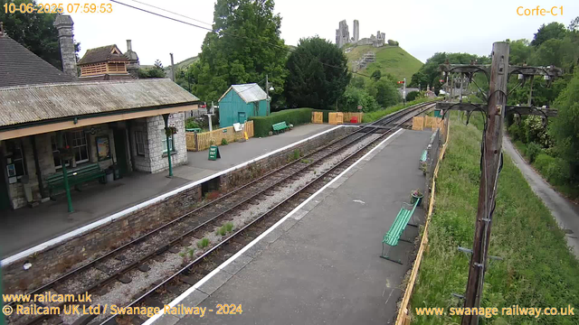 A small railway station platform with a building on the left side that has a sloped, weathered roof and stone walls. There are several green benches along the platform and a sign indicating "Way Out." A green shed is situated behind the benches, and a wooden fence with some flowers and plants borders the platform. In the background, a grassy hill with ruins of an ancient castle is visible, surrounded by lush trees. A single railway track runs through the station, and there are utility poles on the right side of the platform. The sky is overcast, and the timestamp reads 07:59:53 on June 10, 2025.