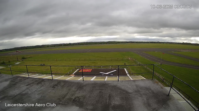 A view of an empty runway and surrounding grassy area at Leicester Aero Club on a cloudy day. The runway is in the distance, with a windsock on a pole to the left and a small helipad marked with a large white "X" in the foreground. Overcast dark gray clouds cover the sky. There are no airplanes or people visible in the image.