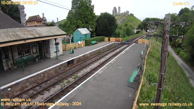 A small railway station platform with two green benches, a stone building with a chimney, and a small turquoise hut in the background. There are yellow safety barriers at the end of the platform and some potted plants on the benches. A railway track runs alongside the platform, with grass and weeds growing between the rails. To the right, there are utility poles with electrical wires, and behind the station, there are lush green trees, a pathway, and a hillside with ruins or a castle at the top. The scene appears to be in a rural area during daytime.