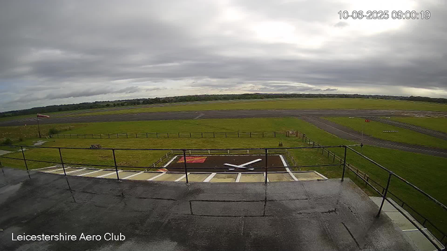A view from a webcam at Leicestershire Aero Club shows an overcast sky with gray clouds, a runway in the background, grassy areas surrounding the runway, and a helicopter landing pad with a white helicopter silhouette on it in the foreground. There is a small flagpole on the left side with a flag, and a person walking along the grassy area on the right side. The timestamp on the image indicates it was taken on October 6, 2025, at 09:00:19.