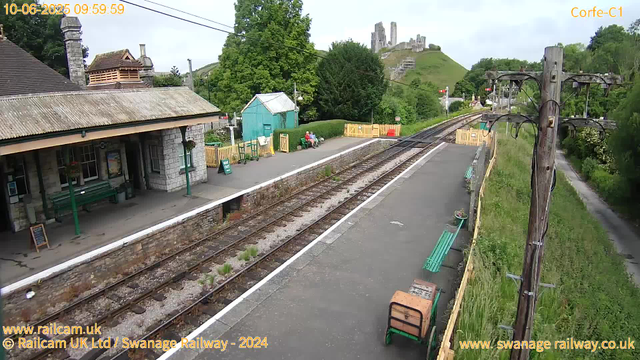 A small railway station platform with a building on the left that has a stone exterior and a tiled roof. There are two green benches, one next to the building and the other further along the platform, where two people are seated. A blue-green shed is situated behind the benches. The railway tracks run through the center of the image, with some vegetation growing between the sleepers. To the right, there are several green benches, a wooden cart, and a row of potted plants along the edge of the platform. A wooden utility pole with wires stands near the platform's edge on the right side. In the background, there are lush green trees, a hill with a castle ruin on top, and some fences at the far end of the station. The sky is partly cloudy, indicating a bright day. The image is time-stamped "10-06-2025 09:59:59".
