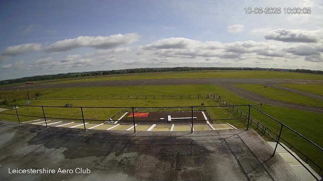 A wide view of an airfield with a tarmac, grassy fields, and a runway stretching into the distance under a partly cloudy sky. The image appears to be taken from a webcam on the Leicestershire Aero Club at 10:00 AM on October 6, 2025. In the foreground, there is a helipad marked with a large white "H" and a small red section. The area has safety barriers and some parking spaces. The horizon shows green fields with trees in the background.