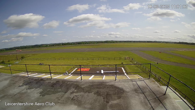 A view from a webcam at Leicestershire Aero Club showing a grassy airfield with runways extending into the distance under a partly cloudy sky. The foreground features a small parking area with designated spaces, a fence, and a windsock on the left side. There are some markings on the ground, and the fields and trees stretch out beyond the airfield. The timestamp in the top right corner indicates the date and time as October 6, 2025, at 11:01 am.