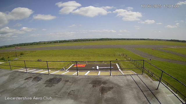 A view from a webcam at Leicestershire Aero Club showing a clear sky with scattered clouds. The scene includes a small helipad with a red and white helicopter landing pad and a white helicopter rotor blade. Beyond the helipad, there is an open grassy area leading to a paved airfield with taxiways and runways in the distance. A windsock can be seen on the left, and a wooden fence borders the grassy area. The timestamp indicates the date and time as October 6, 2025, at 12:01 PM.