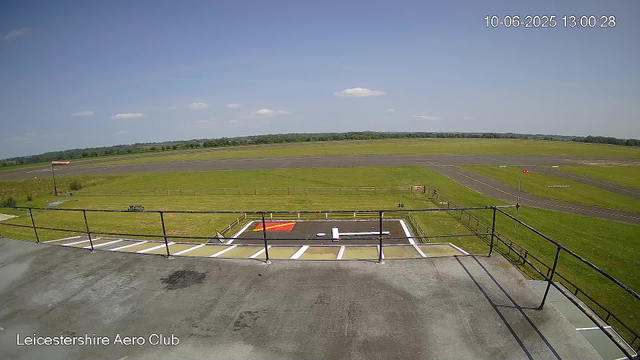 An outdoor view from a webcam at Leicesterhire Aero Club showing a clear, sunny day with a blue sky and a few scattered clouds. In the foreground, there is a rooftop area with safety railings and markings, including a helipad marking with a white "H" and red squares. A grassy area with a flagpole and a small fence is visible beyond the rooftop. In the background, there is a runway with painted markings, surrounded by green fields and trees on the horizon. The timestamp on the image is 10-06-2025 at 13:00:28.