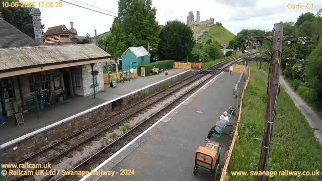 A small railway station with a stone building on the left, a platform with a few people sitting on benches, and a green shed nearby. There are train tracks running through the center of the image. On the right side, a wooden fence and a yellow barrier block the end of the platform. A person is walking along the platform, and another person is sitting on a bench with a small bag beside them. In the background, a lush green hill with an ancient castle ruins is visible under a cloudy sky. There is also a telephone pole with wires running across the scene. The image has a timestamp of June 10th, 2025, at 13:00:29.
