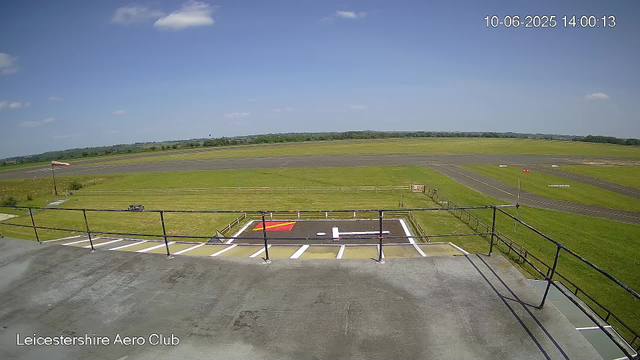 A view from a webcam at Leicestershire Aero Club shows an airfield with a runway stretching into the distance under a partly cloudy sky. In the foreground, there is a helicopter landing pad marked with a large white "H" and surrounding parking spaces, some of which are striped in yellow and white. A fence surrounds the area, and a small windsock is visible on the left side. The camera's timestamp reads 10-06-2025, 14:00:13.