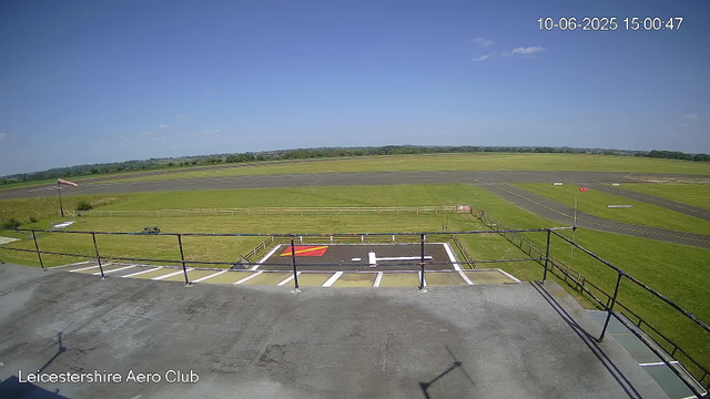 The image shows a view from a webcam at Leicestershire Aero Club. In the foreground, there is a parking area with a designated helicopter landing pad marked with a white "H" and red and yellow markings. Beyond the parking area, the scene features a grassy field enclosed by a small wooden fence, with a windsock on a pole indicating the wind direction. An empty picnic table is also visible near the fence. In the background, there is an open runway and taxiways with a few grass strips, set against a backdrop of green fields and trees under a clear blue sky with a few small clouds. The timestamp in the top right corner shows the date and time as 10-06-2025 at 15:00:47.