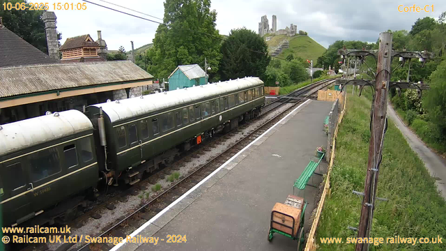 A vintage green train with a white roof is stationary at a small railway platform. Nearby, there are a few green benches, one of which has a brown bag placed on it. To the right, there are wooden utility poles with electrical wires running overhead. In the background, lush green trees, a small teal-colored building, and the ruins of a castle situated on a hill can be seen. The sky is partly cloudy.