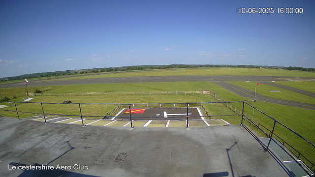 A view from a webcam at Leicestershire Aero Club showing a small helipad with a white and red marking, surrounded by a fence and a parking area. Beyond that, there is a grassy field and an airstrip with runway markings. The sky is clear and blue with a few small clouds. The timestamp in the upper right corner indicates the date and time as October 6, 2025, at 4:00 PM.