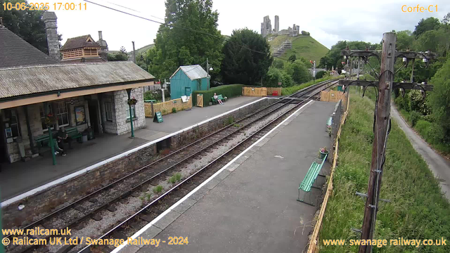 A small train station platform with a stone building on the left side, where a person is sitting on a green bench. There are two green benches on the platform, one of which has a person sitting on it. A yellow fence section separates the platform from the railway tracks, which run through the center of the image. To the right, there are two green benches, one with a flowerpot on it. In the background, there is a green building, trees, and a hill with ruins or castle remains at the top. On the right edge, a tall wooden utility pole with electrical wires is visible, and a narrow path runs alongside the station into a lush green landscape. The scene is overcast with cloudy skies. The timestamp shows the date and time as 10-06-2025 17:00:11.