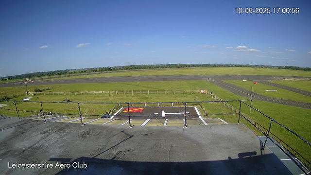 A clear view of an airfield at Leicestershire Aero Club under a blue sky with a few scattered clouds. In the foreground, there is a small helipad with a white H marking and a red and yellow landing pad nearby. The image shows a grassy area enclosed by a fence, with a flagpole flying the Union Jack. The airfield's runway extends into the distance, with markings and signs visible along its length. The timestamp in the top right corner reads "10-06-2025 17:00:56".