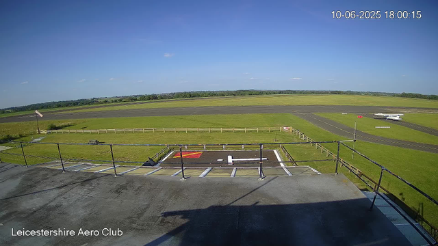 A clear view of a small airfield shows a helicopter parking spot in the foreground, surrounded by a metal railing. Beyond the grassy area, there is a small airplane on the taxiway. The sky is bright and mostly clear with a few small clouds, and the horizon line is visible in the distance. The caption at the bottom left reads "Leicestershire Aero Club." The timestamp in the top right corner indicates the date and time as June 10, 2025, at 6:00 PM.