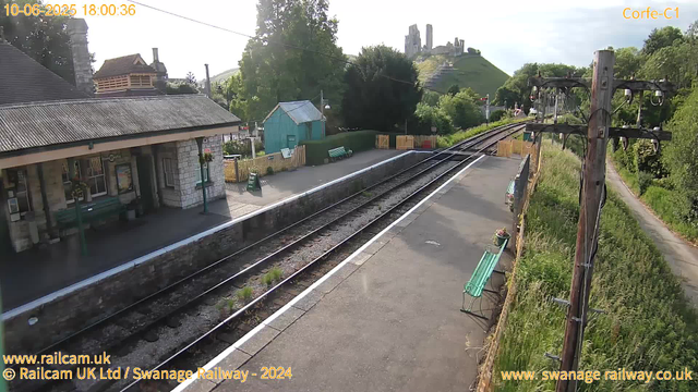 A small railway station platform with a building on the left side, featuring a stone facade and dark roofing. There are green benches and a small blue shed further along the platform. The railway tracks run through the center of the image, with a yellow fence and several plants along the sides. To the right, there are tall wooden utility poles with wires, and a narrow footpath bordering the greenery. In the background, there is a hillside with castle ruins on top, surrounded by trees and lush vegetation. The sky is partly cloudy, and the scene is well-lit by daylight. The timestamp indicates the date and time as June 10, 2025, at 18:00.