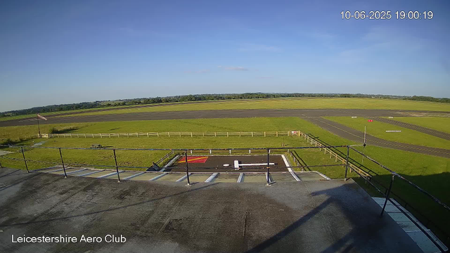 An empty runway at Leicester Aeroclub on a clear day. The sky is blue with a few wispy clouds. In the foreground, there is a rooftop or platform with a railing, a red and white marking, and some equipment. The grass surrounding the runway is green, and there are a few flags and small structures visible. The landscape stretches out with fields and trees in the distance. The timestamp in the top right corner indicates the date and time as October 6, 2025, at 19:00:19.