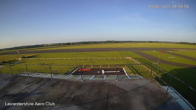 A wide view of an airfield at sunset, with a helipad in the foreground marked with a white "H" and surrounding fencing. The airfield includes grassy areas, taxiways, and runway strips stretching into the distance under a clear blue sky. The timestamp in the upper right corner indicates the date as October 6, 2025, at 7:59 PM. The bottom left corner has the label "Leicestershire Aero Club."