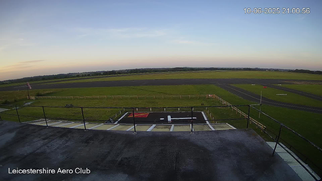 A wide view of an outdoor airfield at sunset, showing a grassy area with a small helipad in the foreground. The helipad has a white helicopter landing pad symbol and red markings. Surrounding the airfield are open fields and a runway in the distance. A wooden fence runs along the grassy area, and there is an American flag on a pole to the left. The sky is clear with a few faint clouds. The date and time in the top right corner are 10-06-2025 21:00:56. The bottom left corner has the text "Leicestershire Aero Club."