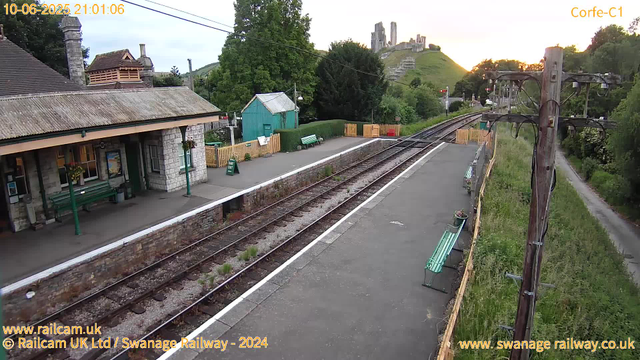 A small train station platform is shown with two green benches, one on each side of the station building. The station building has stone walls, a shingled roof, and an overhang with hanging flower baskets. There is a green shed nearby, surrounded by a yellow picket fence, with a picnic table and some documents attached to it. A small sign indicates the exit. The railway tracks run parallel to the platform, with some weeds growing between the sleepers. To the right, a tall wooden utility pole with electrical wires extends along the platform. In the background, there is a hill with an old castle ruin at the top, and trees surround the area, with the sun setting behind the hill. The timestamp shows the date as June 10, 2025, at 21:01.