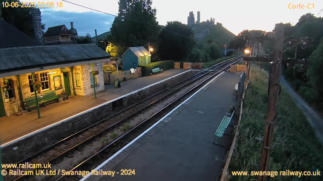 A small railway station platform during twilight hours, with a building on the left that has a green door and hanging flower baskets, and several benches along the platform. The railway tracks run parallel to the platform, stretching into the distance. There are street lamps illuminating the area, and a utility pole with wires is visible on the right side. In the background, there are trees, a small blue shed, and a hill with ruins or old structures silhouetted against the sky, which is beginning to darken. The overall scene appears peaceful and quiet.
