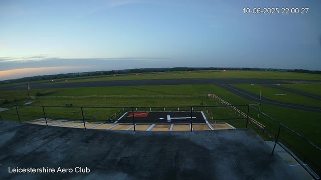 An outdoor view from a webcam at Leicesterhire Aero Club showing a runway and grassy fields under a partly cloudy sky during dusk or dawn. The timestamp indicates the date as October 6, 2025, at 10:00 PM.