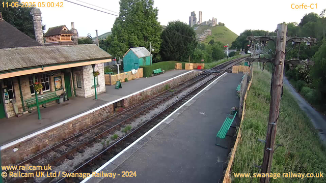 A small train station platform with a stone building on the left, featuring a green door, windows, and hanging flower baskets. There are green benches on the platform, a small green shed, and a "Way Out" sign. The railway tracks run through the center of the image, with a wooden fence separating the platform from the surrounding greenery. Tall tree-covered hills and a castle ruin are visible in the background. On the right, several wooden telephone poles with wires stretch along a grassy embankment, and the sky appears to be early morning or late evening. The image includes timestamps and website credits at the bottom.