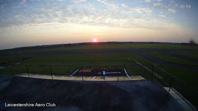 A sunrise over an open grassy area with paved paths, surrounded by a railing, at Leicestershire Aero Club. Some small platforms and equipment are visible in the foreground. The sky is partly cloudy with colors of pink, orange, and blue. The date and time stamp shows 11-06-2025 at 05:00:59.