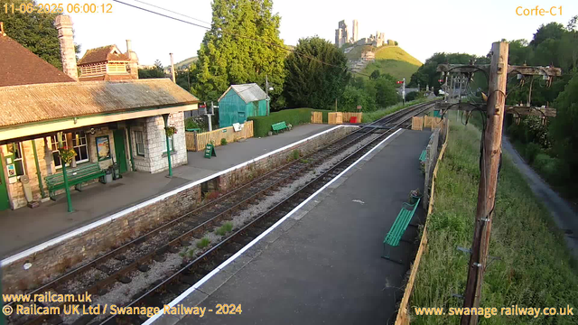 A small railway station platform with a stone building on the left side. There are green benches, a sign that says "WAY OUT," and a green shed in the background. The railway tracks run through the center, curving towards a distant hill with a castle ruin on top. There are large trees and greenery around the station, and an electrical pole with wires is visible on the right side. The scene is bathed in early morning sunlight, creating a warm atmosphere.