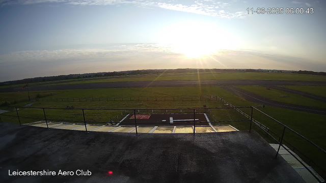 A wide view of an airfield at sunrise, with the sun low on the horizon casting a bright glow across the sky. The foreground shows an observation deck or platform with railings, overlooking green grassy areas and runway strips extending into the distance. The sky is mostly clear with some light clouds. The image is timestamped "11-06-2025 06:00:43" and includes the text "Leicestershire Aero Club" at the bottom left corner.