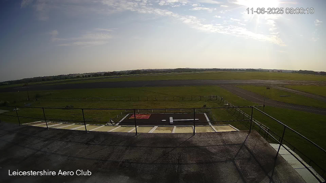 A view from a webcam shows the runway and grassy areas of Leicester Aerospace Club under a partly cloudy sky with sunlight. There is a helipad with a white "H" symbol and surrounding green fields extending into the horizon. The date and time in the top right corner is 11-06-2025, 08:00:10.