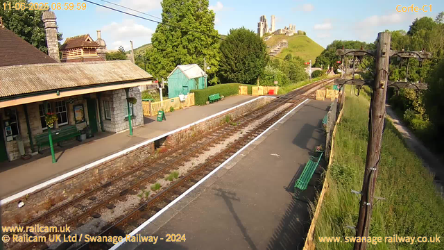 A small train station platform on a sunny day with a stone building and green benches. There are overhead electric wires supported by wooden poles along the tracks. In the background, a hillside with an ancient castle ruins is visible. There are trees and greenery surrounding the area, and a narrow footpath runs alongside the right side of the tracks. The scene appears quiet and peaceful.