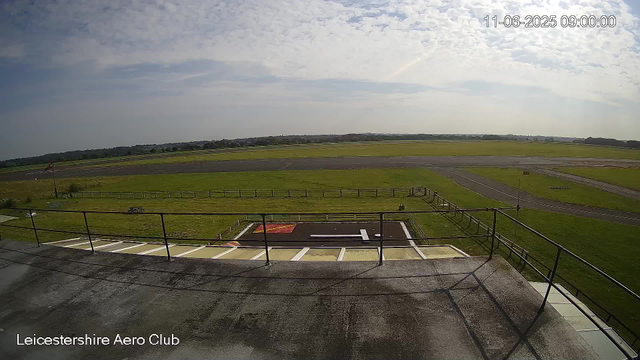A wide view of an airfield showing multiple runways and grassy areas under a partly cloudy sky. In the foreground, there is an observation platform or balcony with a railing, and a helipad marked with an "H" and a red rectangle. The timestamp on the image reads "11-06-2025 09:00:00," and the text "Leicestershire Aero Club" is visible in the bottom left corner.