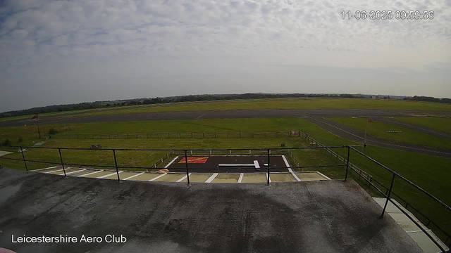 A wide view of an airfield taken from a webcam at the Leicester Shire Aero Club. The foreground shows a helipad marked with a large white "H" and red and white coloring. Beyond the helipad, there is a grassy area with a small bench and a windsock on a pole. The airstrip extends into the distance, with runway markings and grassy fields on either side. The sky is mostly cloudy with patches of sunlight, and a timestamp in the top right corner reads "11-06-2025 09:59:56."