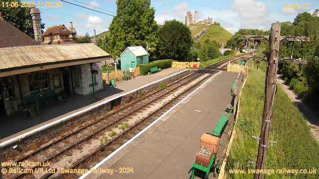 A small train station platform with a building on the left, trees and a green shed in the middle ground, and railway tracks running through the scene. There are benches along the platform, some potted plants, and yellow safety barriers at the far end. In the background, a hill with ancient castle ruins is visible under a partly cloudy sky. A wooden utility pole with wires stands to the right. The scene is bright and sunny.
